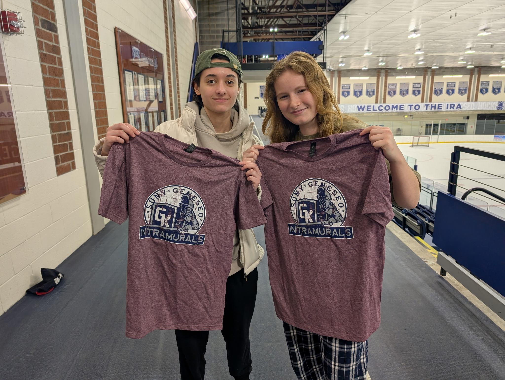 Cornhole champs, 2 students, holding up maroon intramural t shirt, on track above ice rink