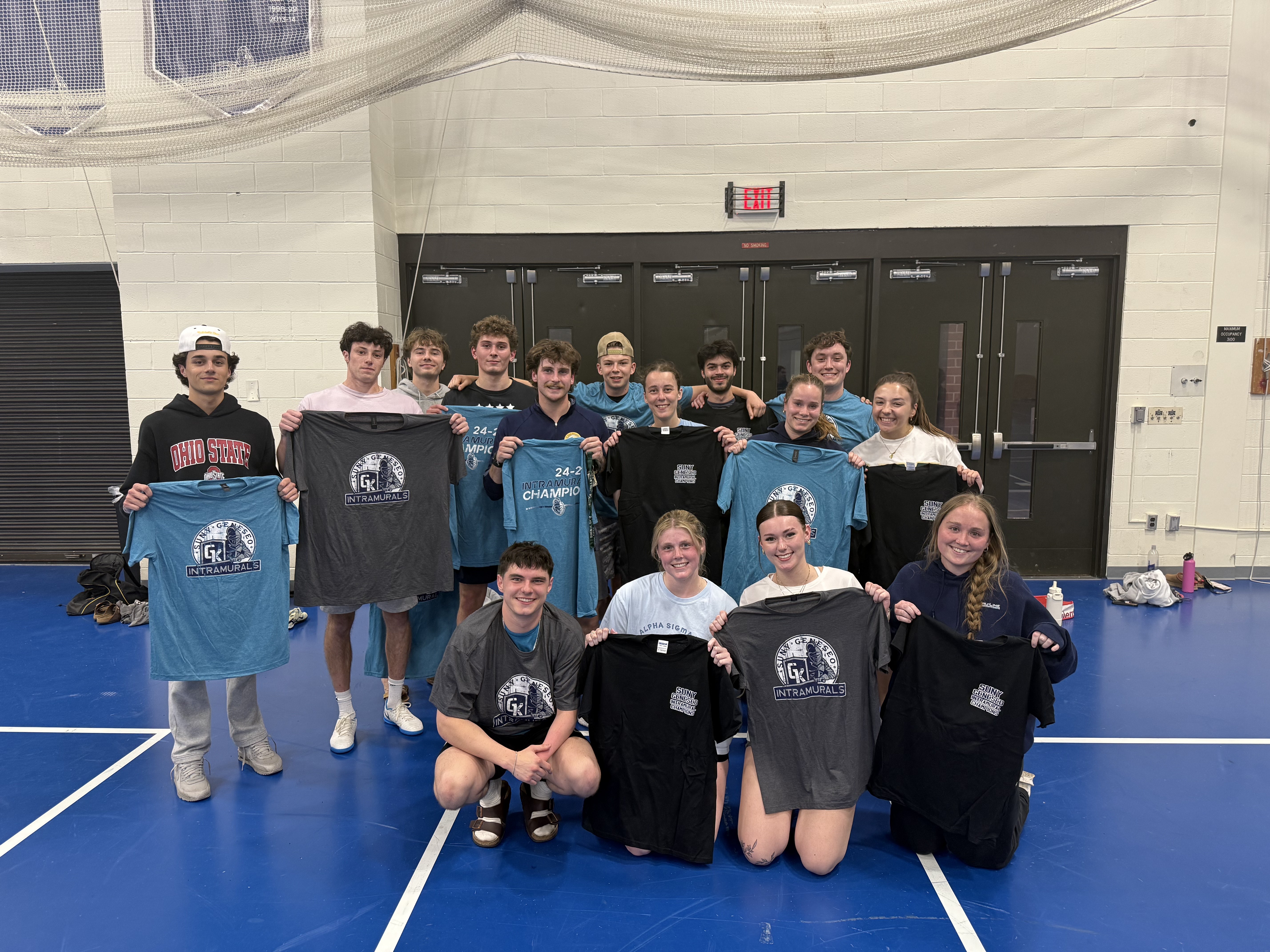group of students in kuhl gym, holding blue, grey, and black champ shirts