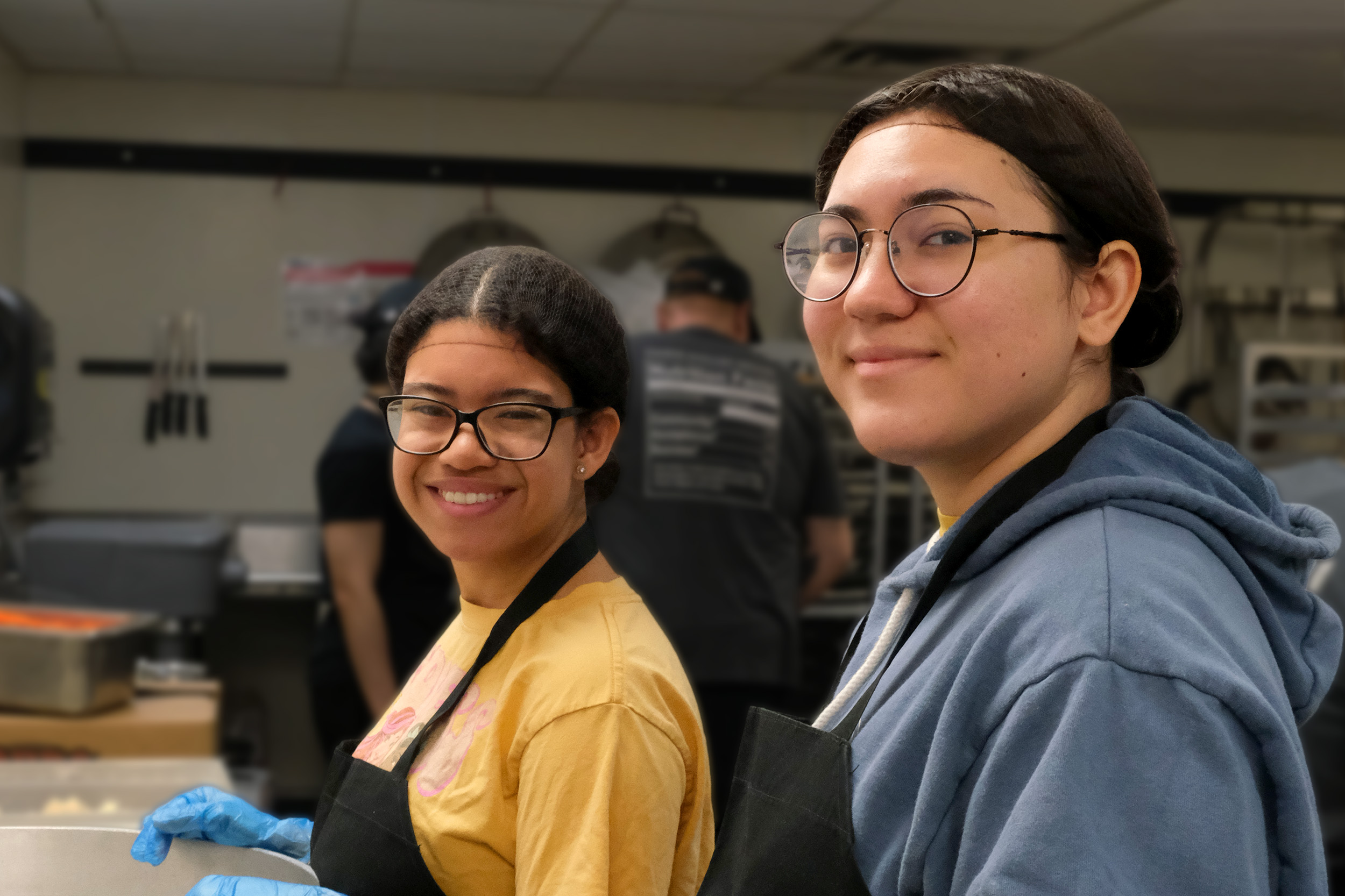 Two students smiling while preparing a meal for a cultural dinner event.