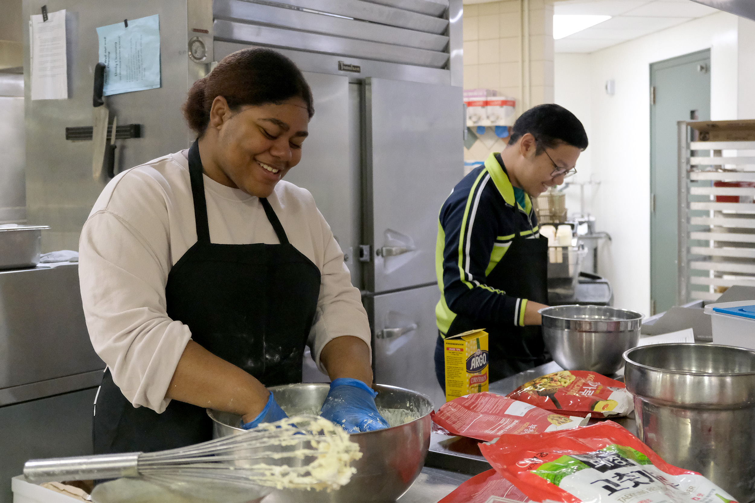 Two students smiling while preparing a meal for a cultural dinner event.