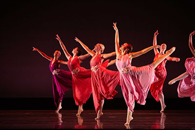 Six dancers in flowing pink and red dresses perform on a dark stage. Captured mid-movement, they extend their arms with one leg raised behind.