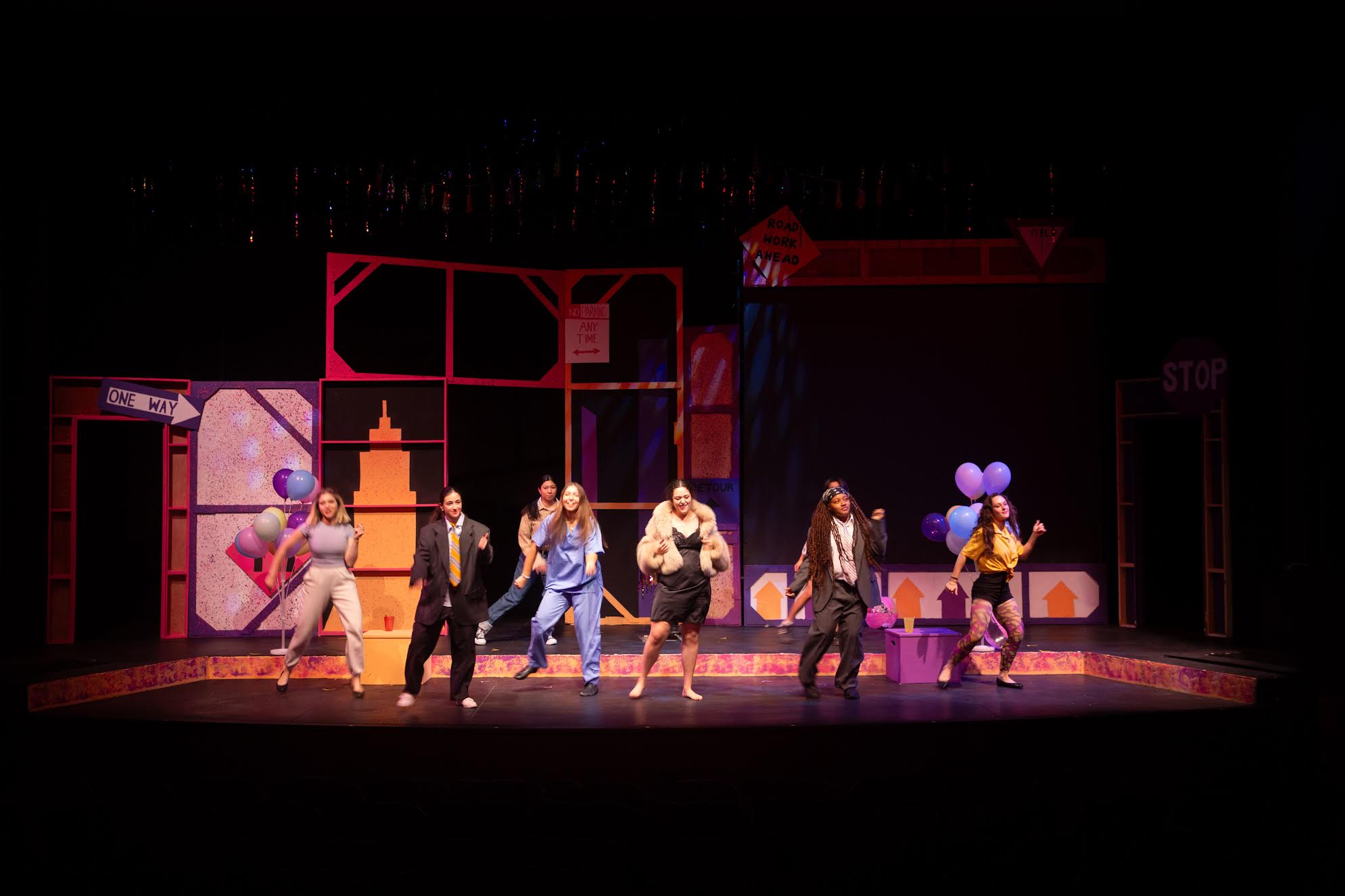 Seven performers on stage in various costumes, set against a backdrop of balloons and signs, including a 'One Way' sign on the left and a 'Stop' sign on the right.