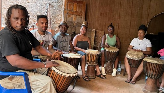 A group of six individuals seated in a semi-circle indoors, each holding a djembe drum. The group appears to be participating in a drumming activity.