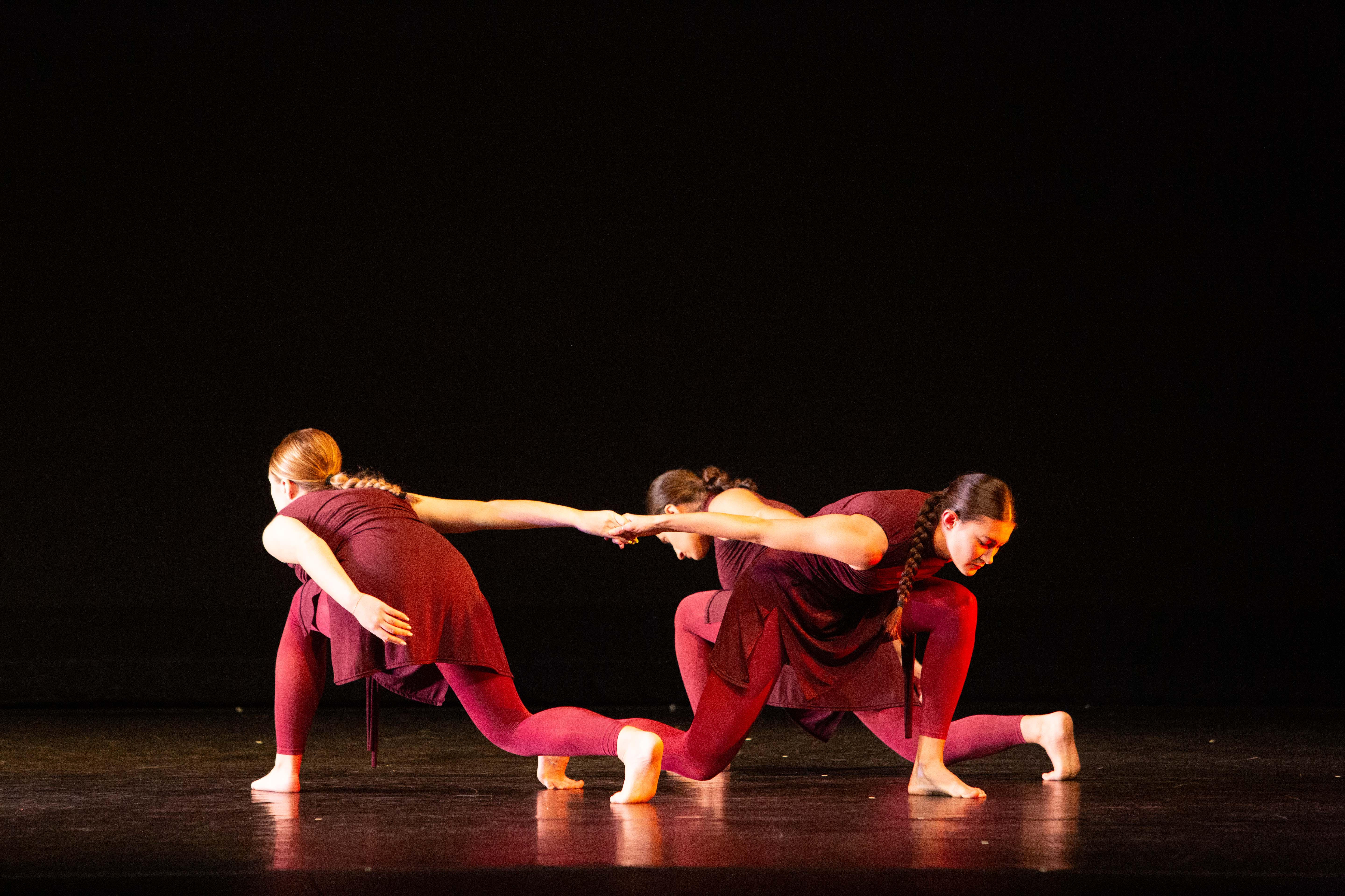 3 dancers in red costumes perform a contemporary routine on stage. One dancer reaches toward another, while the third crouches nearby. The dark background emphasizes their expressive poses and coordinated movement.