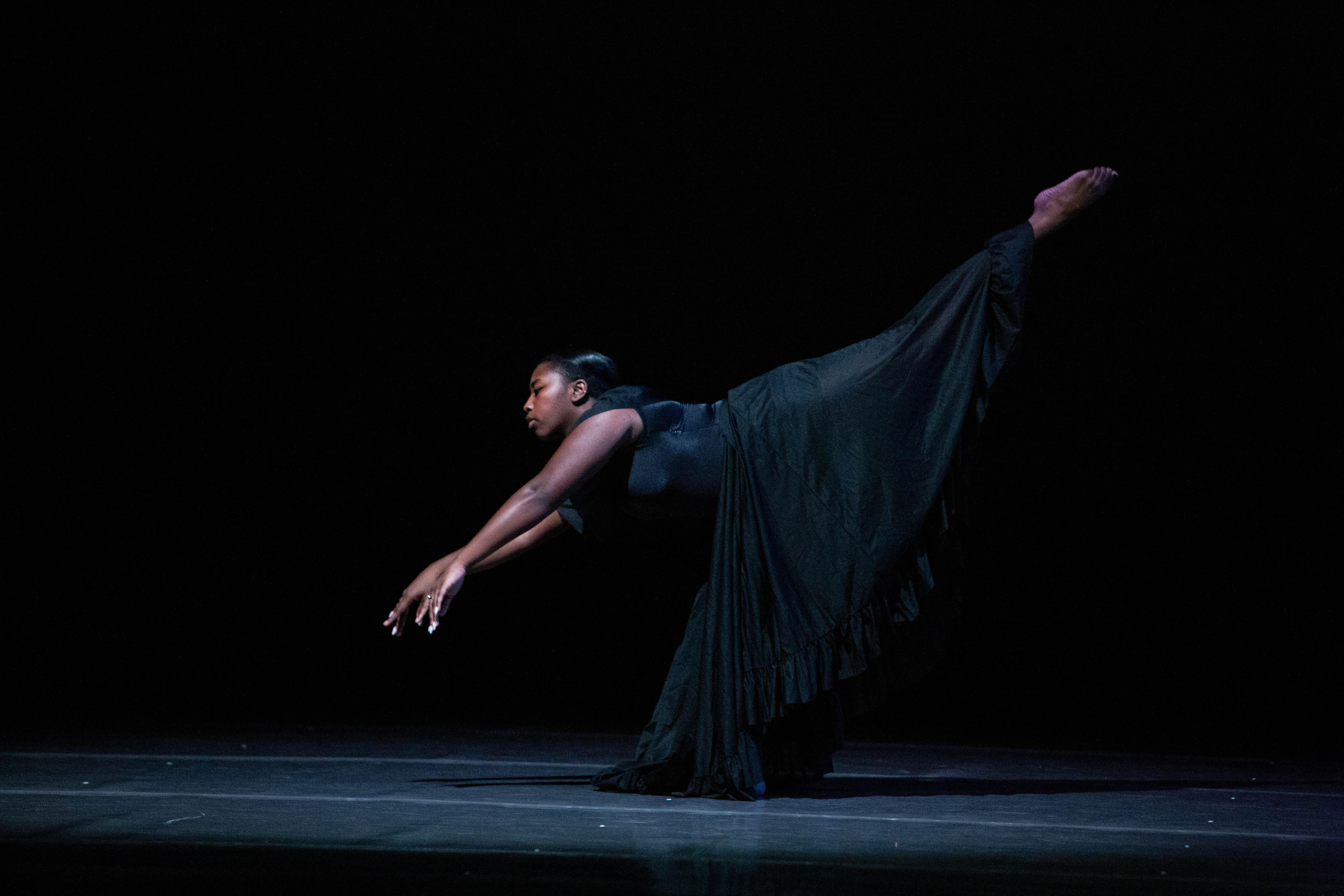 A dancer in a long black dress performs a dramatic move on stage, balancing on one leg with the other extended high behind. Their arms are stretched forward, against a dark background that emphasizes the pose and costume.