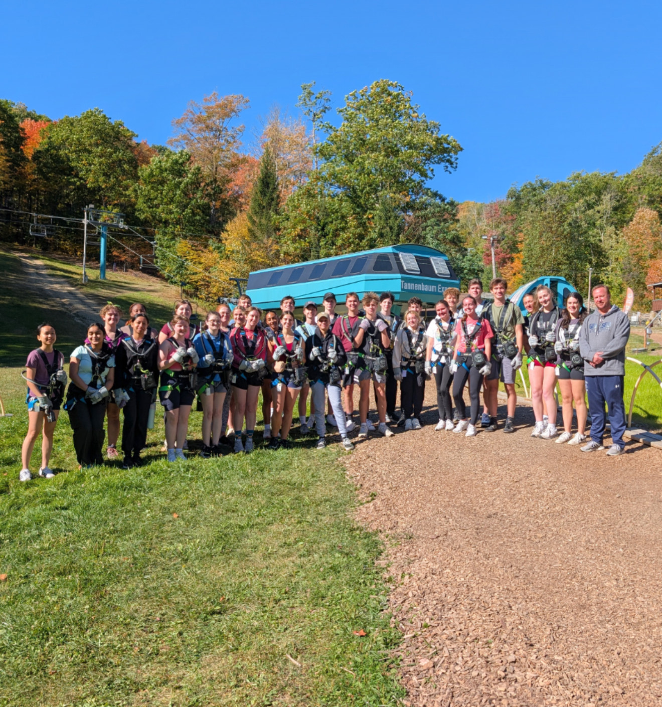A large group of people wearing harnesses pose outdoors in front of a blue gondola labeled 'Tannenbaum Express' on a sunny fall day, with trees in the background.