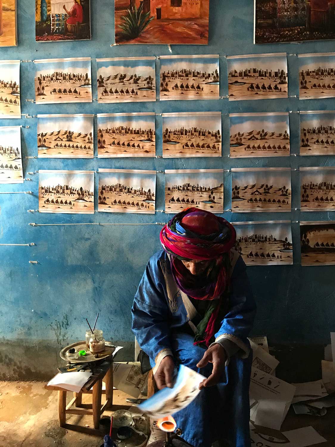 Man in turban painting landscapes on the floor with similar paintings on the wall.