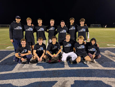 A large group of people on a sports field at night wearing matching gray “SUNY Geneseo Intramurals” shirts, some holding additional shirts, gathered closely for a group photo.