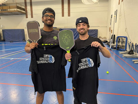 Two students standing on an indoor gym court holding black T-shirts with “SUNY Geneseo Intramurals” printed on them and each holding a pickleball paddle.