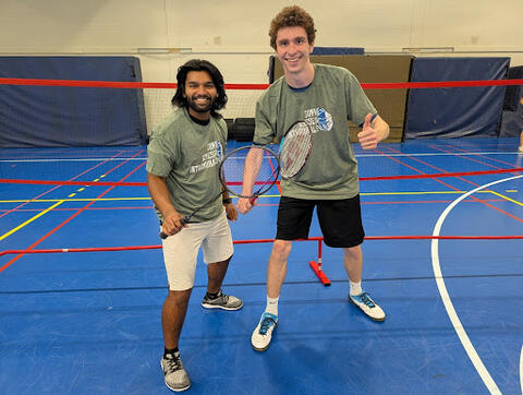 Two students on an indoor badminton court wearing gray “SUNY Geneseo Intramurals” shirts, each holding a badminton racket and standing near a red net.