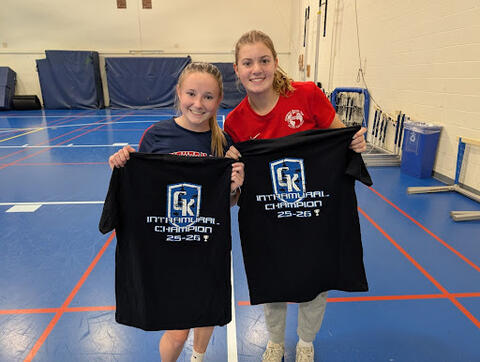 Two students standing on an indoor gym court holding black T-shirts that read “GK Intramural Champion 25-26” in bold blue and white lettering.