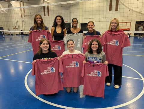 A group of students on an indoor volleyball court holding red and gray “SUNY Geneseo Intramurals” T-shirts, with a volleyball net in the background.