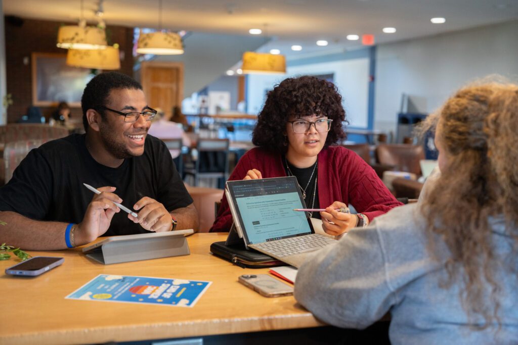 Three SUNY Geneseo students sit together at a table in a campus lounge, studying and collaborating with laptops and notes.