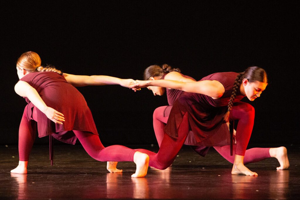unnamed-2 3 dancers in red costumes perform a contemporary routine on stage. One dancer reaches toward another, while the third crouches nearby. The dark background emphasizes their expressive poses and coordinated movement.