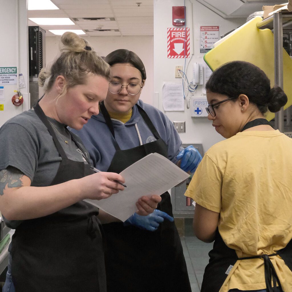 Chef Marissa DioGuardi discusses the cultural dinner menu with two students in the kitchen.